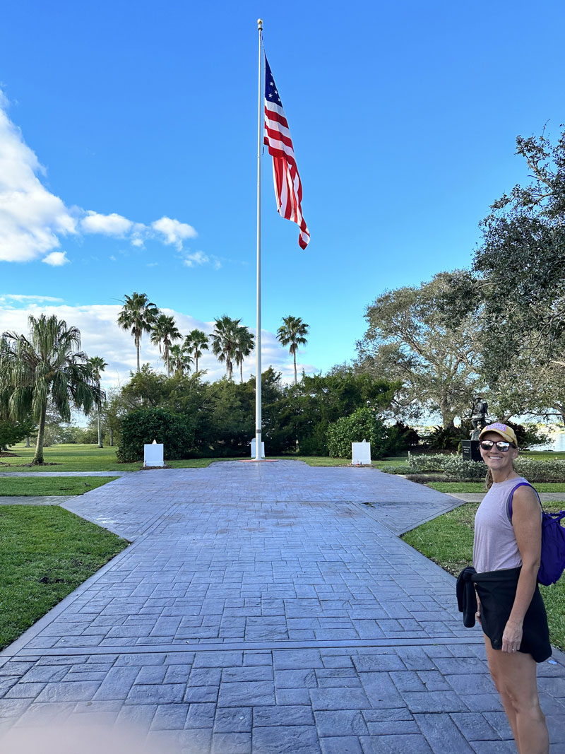 A woman stands in front of a very large American flag with palm trees in the background.